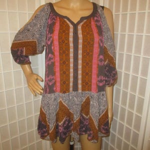May include: A patterned tunic dress with a V-neckline and cut-out shoulders. The dress features a mix of colors including brown, pink, and gray, with a patterned design. The sleeves are three-quarter length.