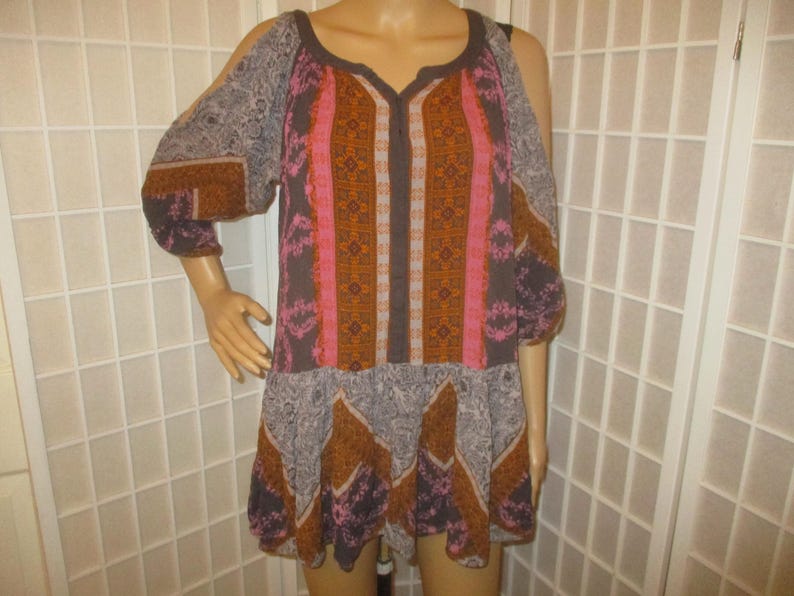 May include: A patterned tunic dress with a V-neckline and cut-out shoulders. The dress features a mix of colors including brown, pink, and gray, with a patterned design. The sleeves are three-quarter length.