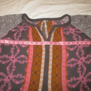 May include: A patterned top with a V-neck and long sleeves. The top features a combination of colors, including gray, pink, and orange. A pink measuring tape is laid across the top, measuring approximately 22 inches across.