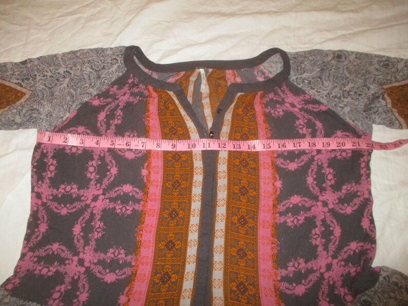 May include: A patterned top with a V-neck and long sleeves. The top features a combination of colors, including gray, pink, and orange. A pink measuring tape is laid across the top, measuring approximately 22 inches across.