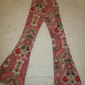 Puede incluir: Pantalones acampanados con un vibrante estampado de cachemira en tonos rojos, rosas, marrones y crema. Los pantalones est&aacute;n extendidos, mostrando toda la longitud y el intrincado dise&ntilde;o. La tela parece suave y ligera.