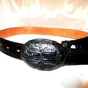 May include: Black leather belt with a large, oval buckle. The buckle features a detailed embossed design. The belt has a brown interior and a textured, crocodile-like exterior. A classic accessory.
