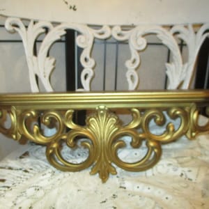 May include: A gold-colored decorative shelf with ornate scrollwork. The shelf is made of metal and has a rectangular shape.