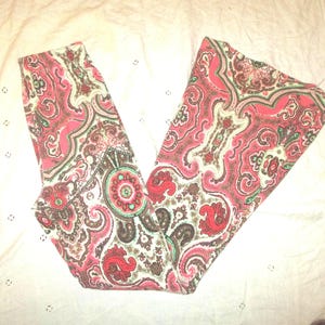 Puede incluir: Pantalones con estampado de cachemira en tonos rojos, rosas, marrones y blancos. La tela presenta un dise&ntilde;o detallado y arremolinado con elementos florales y abstractos. Los pantalones est&aacute;n doblados, mostrando el patr&oacute;n completo.