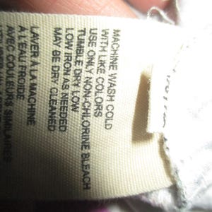 Puede incluir: Primer plano de una etiqueta de tela con instrucciones de cuidado. La etiqueta beige tiene texto negro, incluyendo "MACHINE WASH COLD", "TUMBLE DRY LOW" y "MAY BE DRY CLEANED". La etiqueta est&aacute; parcialmente sujeta por una mano.