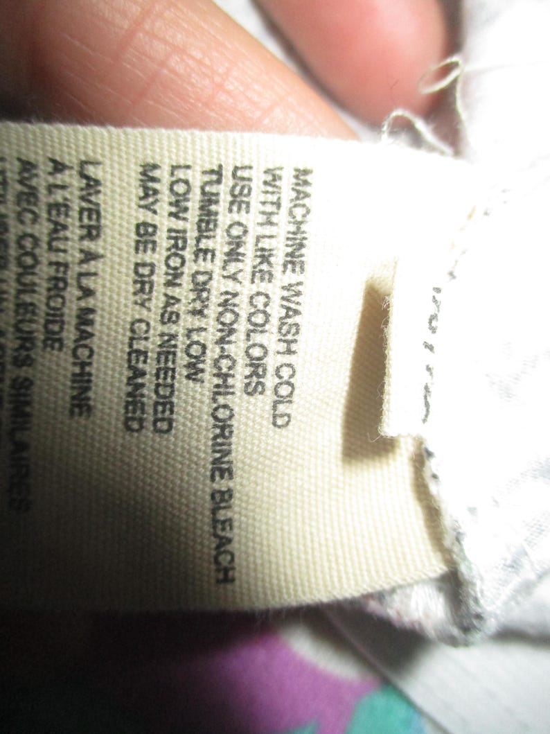 Puede incluir: Primer plano de una etiqueta de tela con instrucciones de cuidado. La etiqueta beige tiene texto negro, incluyendo "MACHINE WASH COLD", "TUMBLE DRY LOW" y "MAY BE DRY CLEANED". La etiqueta est&aacute; parcialmente sujeta por una mano.