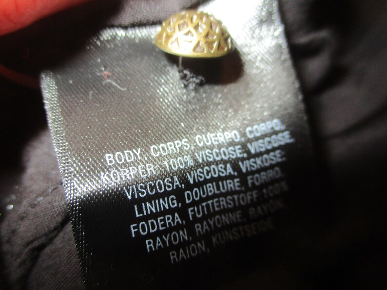 May include: A black fabric label with white text detailing the fabric composition of the garment. The text reads: "BODY, CORPS, CUERPO, CORPO KORPER: 100% VISCOSE, VISCOSE VISCOSA, VISCOSA, VISKOSE LINING, DOUBLURE, FORRO FODERA, FUTTERSTOFF 100% RAYON, RAYONNE, RAYON RAION, KUNSTSEIDE"