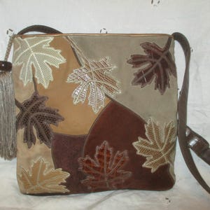 May include: A square shoulder bag in shades of brown and tan featuring a patchwork design with embroidered maple leaf appliqués in various textures. A long, light brown tassel with a dark bead hangs from the side, and a brown strap is attached.
