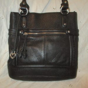 May include: Black leather tote bag with silver-tone hardware. Features a zipped top, front pocket, and decorative tassels. The bag has two top handles and a heart-shaped charm.