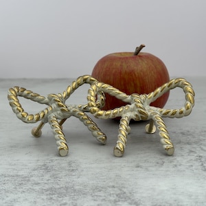May include: Two decorative bow-shaped objects with a white and gold rope design. The bows are made of metal and have three legs. A red apple with a brown stem is behind the bows.