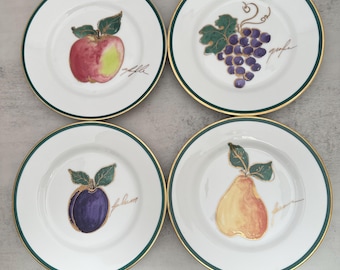 Può includere: Un set di quattro piatti in ceramica bianca con bordo verde scuro e oro. Ogni piatto presenta un'illustrazione in stile acquerello di un frutto diverso: una mela, un grappolo d'uva, una prugna e una pera, con testo in corsivo.
