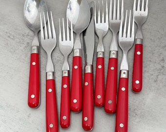 Può includere: Un set di posate in acciaio inossidabile con manici rosso brillante e rivetti argentati. La collezione comprende due cucchiai, un coltello e cinque forchette, tutti disposti insieme. Ogni pezzo ha una testa in metallo lucido e un manico rosso.