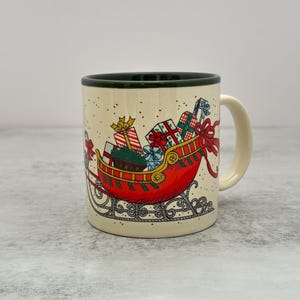 May include: A cream-colored ceramic mug with a dark green rim and handle. The exterior features a festive illustration of a red sleigh filled with wrapped gift boxes, tied with red ribbons, and decorated with small black dots resembling falling snow.