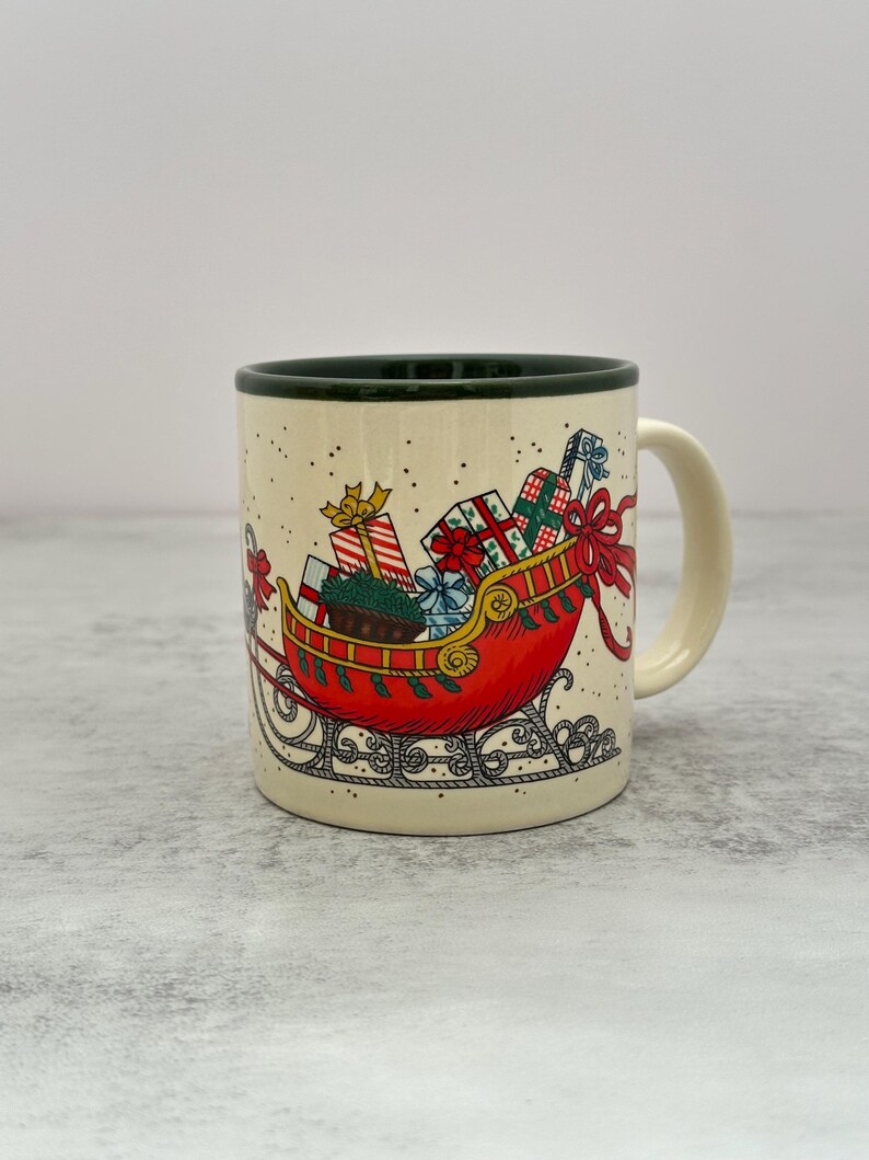 May include: A cream-colored ceramic mug with a dark green rim and handle. The exterior features a festive illustration of a red sleigh filled with wrapped gift boxes, tied with red ribbons, and decorated with small black dots resembling falling snow.
