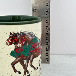 May include: A cream-colored ceramic mug with a dark green rim, featuring a graphic of two brown horses wearing red bridles and green holiday wreaths. A metal ruler is positioned vertically, showing the mug height is approximately 4.5 inches.