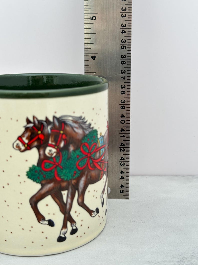 May include: A cream-colored ceramic mug with a dark green rim, featuring a graphic of two brown horses wearing red bridles and green holiday wreaths. A metal ruler is positioned vertically, showing the mug height is approximately 4.5 inches.