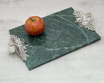 Può includere: Un vassoio rettangolare in marmo verde scuro con due manici in metallo argentato a forma di grappoli d'uva e foglie. Una mela rossa e gialla poggia sulla superficie liscia e venata del vassoio in pietra, mettendone in risalto il contrasto.