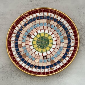May include: A circular decorative plate featuring a mosaic pattern of small square tiles in concentric rings. The colors include deep red, navy blue, light blue, beige, white, yellow, and green, with gold-toned accents and a gold rim.