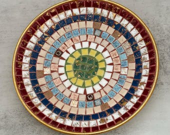 Può includere: Un piatto decorativo circolare caratterizzato da un motivo a mosaico di piccole tessere quadrate disposte in cerchi concentrici. I colori includono rosso scuro, blu navy, azzurro, beige, bianco, giallo e verde, con accenti dorati e bordo.