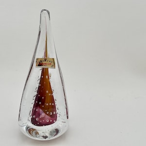 May include: A clear glass teardrop-shaped vase with a brown and purple interior. The vase has small air bubbles throughout and a label that reads "CRYSTAL STYRCAR". The vase is on a white background.