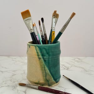 May include: A ceramic art brush holder with a teal and beige glaze, filled with various paint brushes. Several brushes are outside the holder. The brushes have wooden handles and different brush head shapes and sizes.