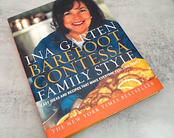 Può includere: Un libro di cucina con copertina rigida intitolato Ina Garten Barefoot Contessa Family Style. La copertina presenta una fotografia di una persona sorridente con una camicia blu, con il testo Easy Ideas and Recipes That Make Everyone Feel Like Family e The New York Times Bestseller, insieme a un'immagine di cibo cucinato con fette di limone.