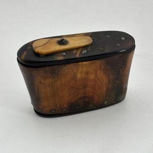 May include: A small, oval-shaped wooden box with a hinged lid. The box is made of dark wood with a light wood inlay. The lid has a small, round knob.