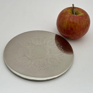 May include: A round, silver-colored metal plate with an ornate, etched floral design. A red and yellow apple with a green stem sits next to the plate. The plate is likely a decorative serving piece.
