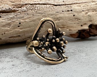 Può includere: Un anello in bronzo unico con un design astratto. L'anello presenta un gruppo centrale di piccoli accenti sferici e una fascia traforata curva. Il metallo ha una finitura scura ossidata con riflessi dorati.