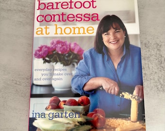 Può includere: Un libro di cucina con copertina rigida intitolato Barefoot Contessa at Home di Ina Garten. La copertina mostra una persona con una camicia blu che taglia del mais su un tagliere di legno, con una ciotola di pesche e un vaso di peonie rosa sullo sfondo. Il testo recita everyday recipes you'll make over and over again.