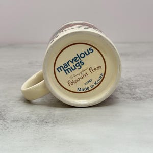 May include: The underside of a cream-colored ceramic mug features blue and brown text reading Marvelous Mugs, The Company of Choice!, Potpourri Press, 1987, and Made in Korea. The mug has a curved handle extending from the side of the base.
