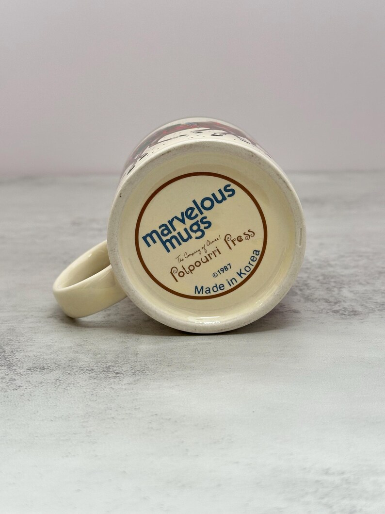 May include: The underside of a cream-colored ceramic mug features blue and brown text reading Marvelous Mugs, The Company of Choice!, Potpourri Press, 1987, and Made in Korea. The mug has a curved handle extending from the side of the base.