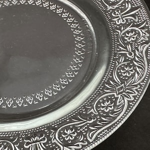 May include: Clear glass plate with an ornate, embossed floral and scroll design around the rim. The plate features a detailed pattern of flowers, leaves, and geometric shapes, creating a vintage aesthetic.