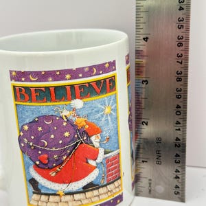 Puede incluir: Taza de cer&aacute;mica blanca con una ilustraci&oacute;n festiva de Pap&aacute; Noel llevando un saco de juguetes en un tejado, con la palabra "BELIEVE" encima. La taza mide aproximadamente 10 cm de alto.