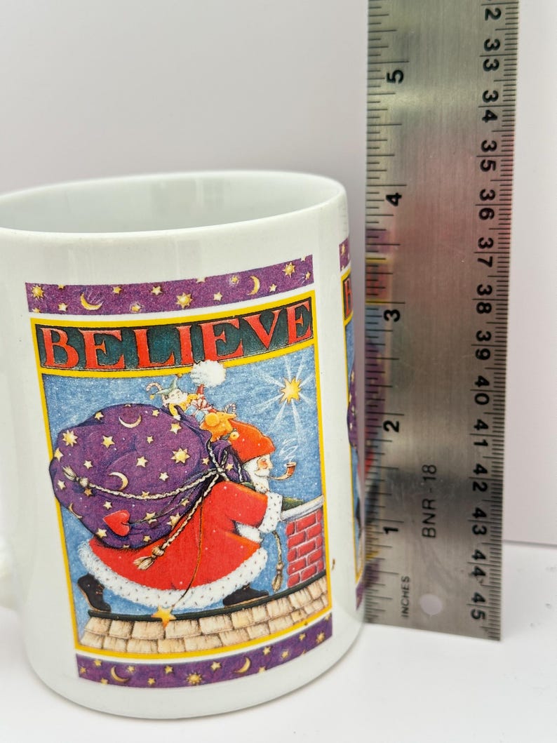 Puede incluir: Taza de cer&aacute;mica blanca con una ilustraci&oacute;n festiva de Pap&aacute; Noel llevando un saco de juguetes en un tejado, con la palabra "BELIEVE" encima. La taza mide aproximadamente 10 cm de alto.