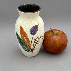 May include: A small white ceramic vase with a brown rim and a floral design in shades of orange, green, and purple. The vase is sitting on a gray surface next to a red apple.