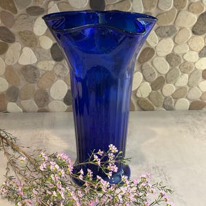 May include: A tall, cobalt blue glass vase with a ruffled rim. The vase has a flared shape and a ribbed texture. A sprig of pink flowers rests in front of the vase, adding a touch of color and contrast.