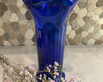 Può includere: Un vaso alto di vetro blu cobalto con bordo a balze. Il vaso ha una forma svasata e una texture a coste. Un rametto di fiori rosa è posto davanti al vaso, aggiungendo un tocco di colore e contrasto.