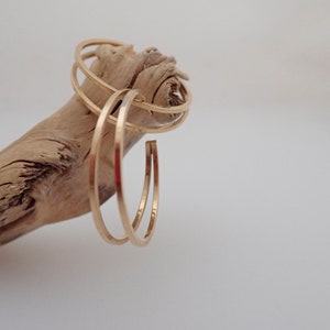 May include: Three gold hoop earrings of varying sizes, stacked on a piece of driftwood.