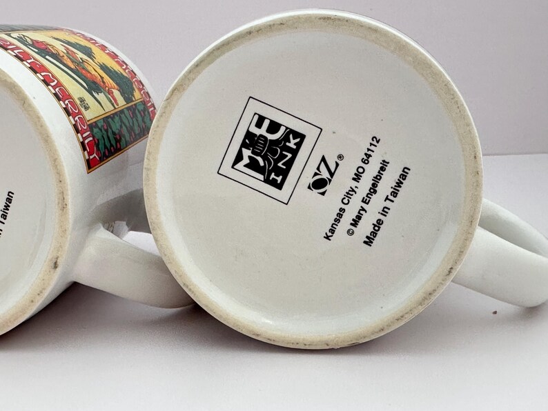 Puede incluir: Dos tazas de cer&aacute;mica blanca con asas, una con una ilustraci&oacute;n colorida y la otra con un logotipo y texto. La base de una taza muestra el texto "Made in Taiwan". Las tazas est&aacute;n sobre una superficie blanca.