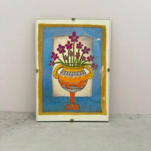 May include: Framed artwork featuring a vibrant orange vase with purple flowers. The vase has decorative handles and sits on a red pedestal. The background is blue with a gold border, all encased in a white frame.