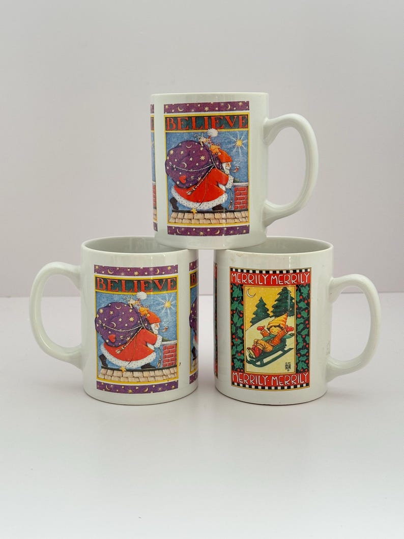 Puede incluir: Tres tazas de cer&aacute;mica blanca con dise&ntilde;os navide&ntilde;os. Dos tazas presentan a Pap&aacute; Noel con la palabra "BELIEVE". La tercera taza muestra a un ni&ntilde;o esquiando con las palabras "MERRILY MERRILY".