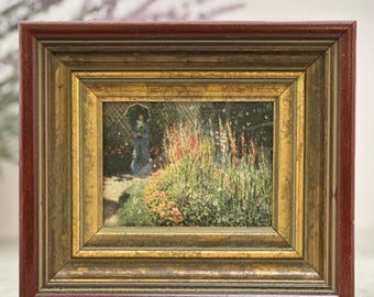 Può includere: Una cornice rettangolare con un bordo esterno marrone rossiccio scuro e una finitura interna dorata anticata. La cornice contiene una stampa di una scena di giardino con una persona che tiene un ombrello verde tra fiori alti.