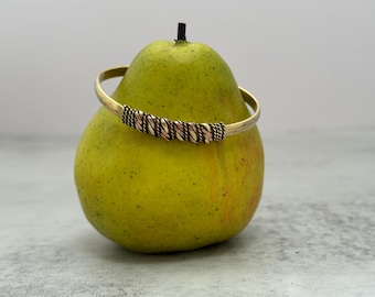 Può includere: Un braccialetto in metallo dorato con un design intrecciato al centro, appoggiato su una pera verde. Il braccialetto ha un design semplice ed elegante. La pera è giallo-verde con un gambo marrone.