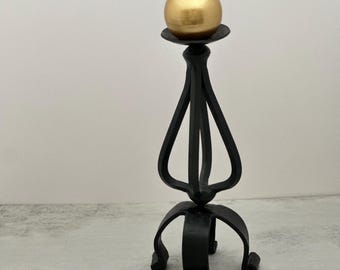 Può includere: Un portacandele in metallo nero con stelo curvo a telaio aperto e base a treppiede, sormontato da una candela sferica dorata. Il portacandele presenta un piattino circolare sulla sommità per sostenere la candela rotonda con stoppino.