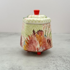 May include: A hexagonal ceramic jar with a lid, featuring a vibrant botanical print of protea flowers in shades of pink, orange, and green. The jar has a small red knob on the lid and stands on three small red feet, with a light green lattice pattern.