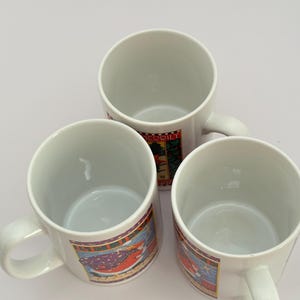 Puede incluir: Tres tazas de cer&aacute;mica blanca con asas, cada una con una banda decorativa colorida alrededor del centro. Las bandas muestran un dise&ntilde;o festivo con una combinaci&oacute;n de colores rojo y verde. Las tazas est&aacute;n vac&iacute;as y dispuestas sobre una superficie blanca.