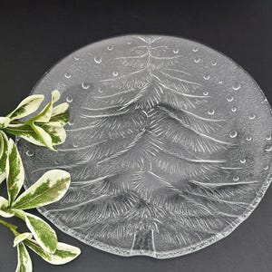 May include: Clear glass plate with an embossed Christmas tree design. The plate features a detailed tree pattern in the center, surrounded by small, embossed circles. A sprig of green and white leaves is in the corner.