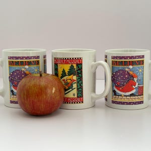 Puede incluir: Tres tazas de cer&aacute;mica blanca con dise&ntilde;os navide&ntilde;os. Dos tazas presentan a Pap&aacute; Noel con un saco, la palabra "BELIEVE" y una combinaci&oacute;n de colores morado y dorado. La tercera taza tiene un esquiador y las palabras "MERILY MERRY". Una manzana est&aacute; delante.