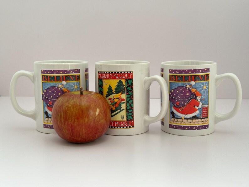 Puede incluir: Tres tazas de cer&aacute;mica blanca con dise&ntilde;os navide&ntilde;os. Dos tazas presentan a Pap&aacute; Noel con un saco, la palabra "BELIEVE" y una combinaci&oacute;n de colores morado y dorado. La tercera taza tiene un esquiador y las palabras "MERILY MERRY". Una manzana est&aacute; delante.